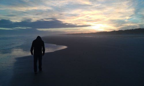 Rear view of man wearing hooded shirt at beach against sky during sunset