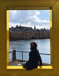 Woman sitting by railing against river and buildings