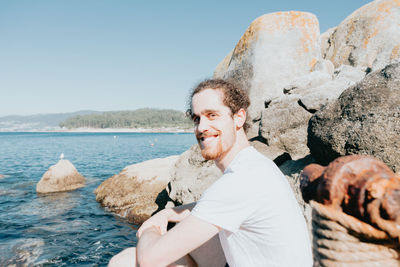 Portrait of man sitting on rock by sea against sky