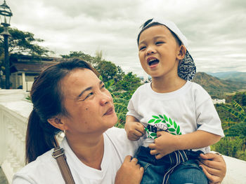 Mother looking at cute son crying while sitting on retaining wall against cloudy sky