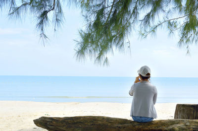 Rear view of woman sitting on fallen tree trunk at beach against clear sky