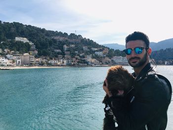 Portrait of man holding dog standing against sea