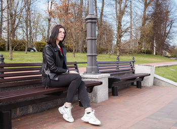 Full length portrait of young woman sitting on bench in park