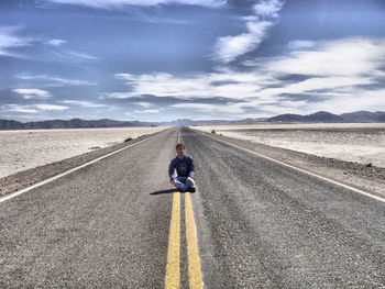 Man sitting on road leading towards mountains against blue sky