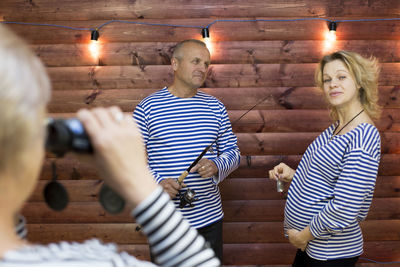 Woman clicking picture of couple with fishing rod while standing against wooden wall