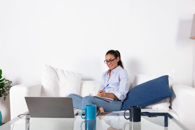Young adult woman taking notes with her laptop on the couch in living room at home with copy space.
