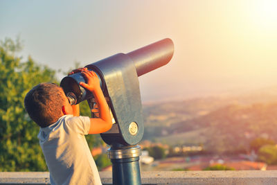 Boy looking through coin-operated binoculars against sky during sunset