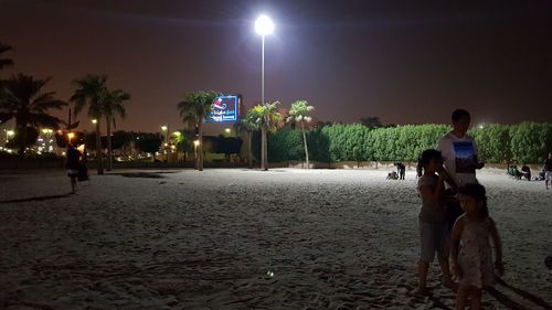 People sitting on illuminated street against sky at night