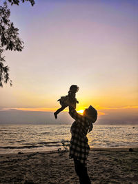 Mother lifting daughter at beach against sky during sunset
