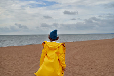 Back view of woman in bright yellow cloak and red hat on shore of north sea 