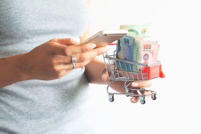 Midsection of woman holding currency in miniature shopping cart against white background