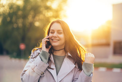 Smiling beautiful girl with long hair in a grey trench coat using smartphone in the spring 