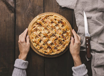 Two female hands hold a round baked pie with apple filling on a wooden board, brown table. view from 