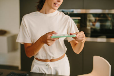 Young female blogger taking photo of healthy vegetable salad in bowl placed on table in kitchen while using smartphone