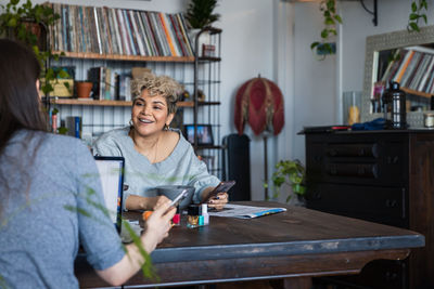 Smiling woman with smart phone looking at friend while sitting in living room