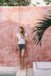 Young woman standing in front of a coral coloured wall touching her hair in tulum, mexico