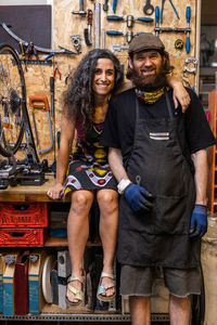 Positive ethnic couple of owners of bicycle service workshop looking at camera and smiling friendly against weathered wall with various repair instruments