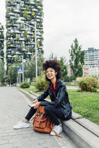 Happy young woman sitting with mobile phone and backpack at park
