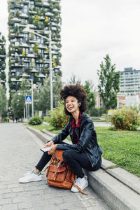 Happy young woman sitting with mobile phone and backpack at park