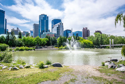 Summer view of fountain on a river against trees, city buildings and blue sky with clouds.