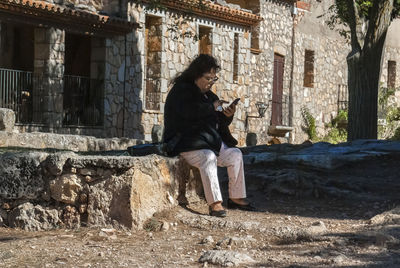 Woman using mobile phone while sitting against building