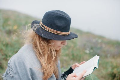 Young woman in hat reading book on field against sky