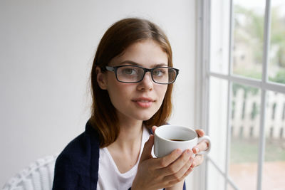 Young woman holding coffee cup while standing against window