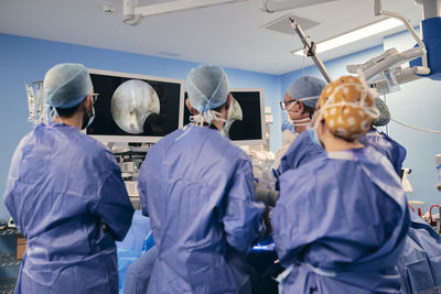 Professionals wearing protective face mask monitoring surgery on computer screen while standing at operating room during covid-19