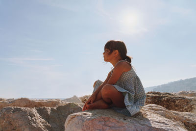 Side view of 6 years girl sitting on rock against sky with closed eyes