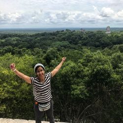 Woman with arms raised standing by tree against sky