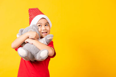 Cute boy holding stuffed toy standing against yellow background