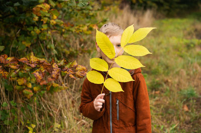 A boy stands on the street and holds a yellow branch with leaves in front of his face