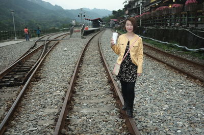 Full length portrait of woman holding disposable cup while standing on railroad track