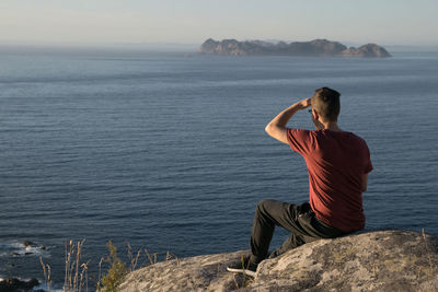 Rear view of man sitting on rock by sea against sky