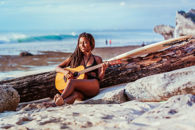 Woman playing guitar while sitting at beach against sky