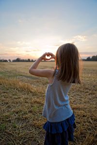 Rear view of girl making heart shape with hands while standing on field against sky