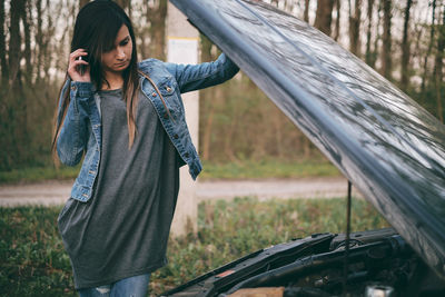 Young woman talking on mobile phone while looking at engine of car