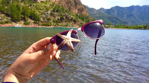Close-up of cropped hand holding sunglasses with dead starfish by lake