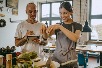 Happy female chef sprinkling chopped vegetable in pan while standing by colleague with diary at studio