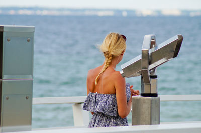 Woman standing by coin-operated binoculars on pier against sea