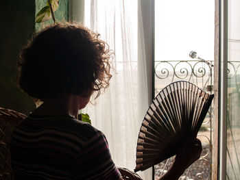 Woman holding hand fan while sitting on seat at home