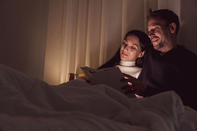 Smiling couple reading book while sitting on bed at home