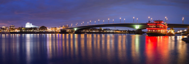 Illuminated bridge over river by buildings against sky at night