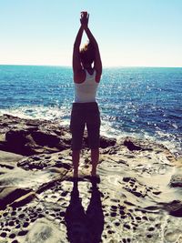 Rear view full length of woman with arms raised against sea on rock during sunny day