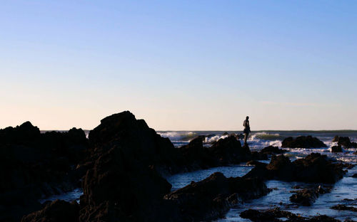 Silhouette man standing on rock by sea against clear sky during sunset