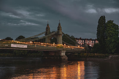 Illuminated bridge over river against cloudy sky