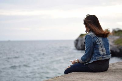 Side view of woman looking at sea while sitting on retaining wall against sky