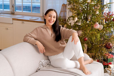 Happy woman sits on couch in sweater next to natural christmas tree, holiday