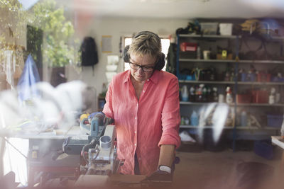 Senior woman using circular saw in workshop seen through glass window