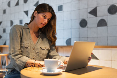 Young focused female browsing internet on portable computer at table with hot drink in cafeteria